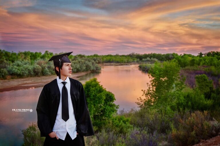 high school senior graduate in cap and gown standing by the rio grande river at sunset in albuquerque